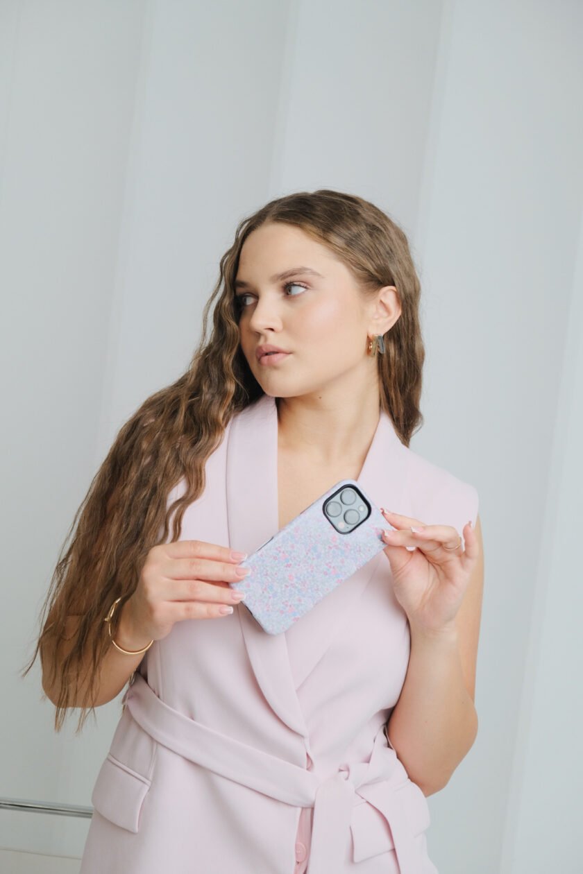 A phone case with a soft floral pattern featuring pink, purple, and blue flowers, placed on a white surface surrounded by pastel-colored sticky notes, colored pencils, and a purple-lined notebook.